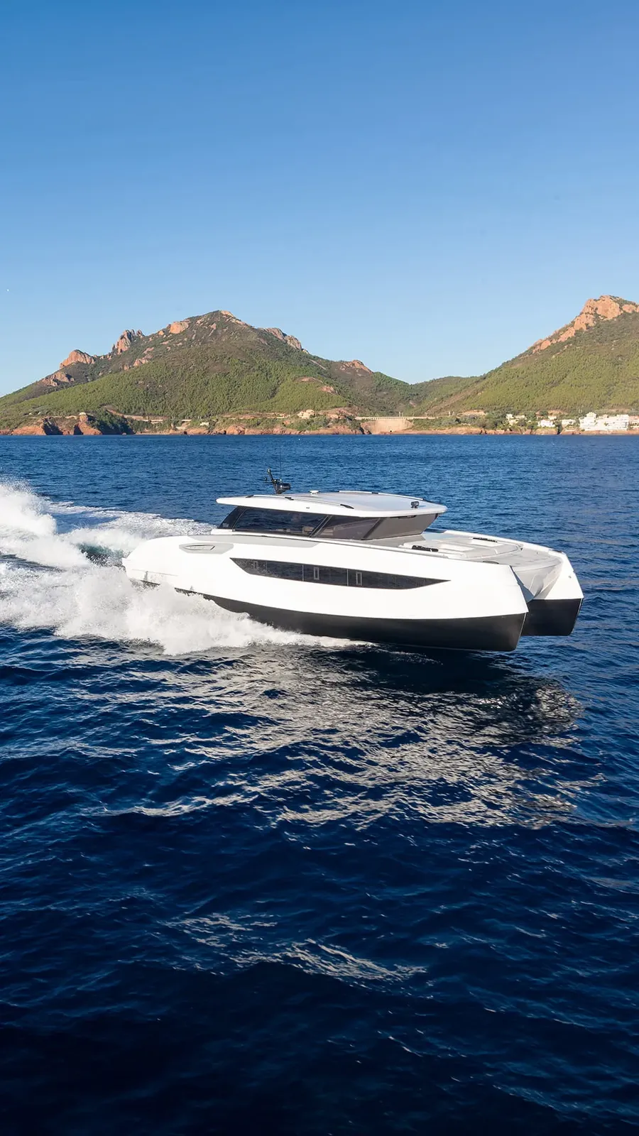 A Sunreef Yachts catamaran cruises over calm seas, with a scenic mountain backdrop under clear skies.