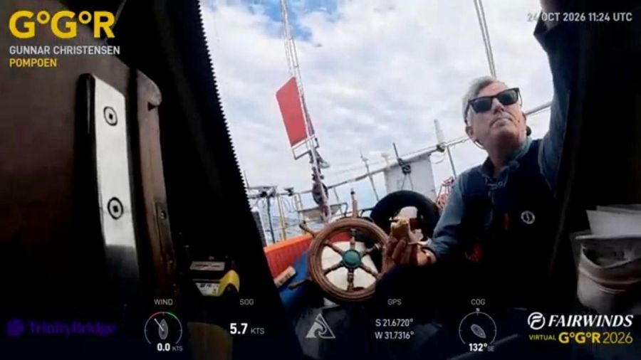 Gunnar Christensen in sunglasses and a lifejacket on the deck of Pompoen, one arm raised, with the ship's wheel and a blue sky behind
