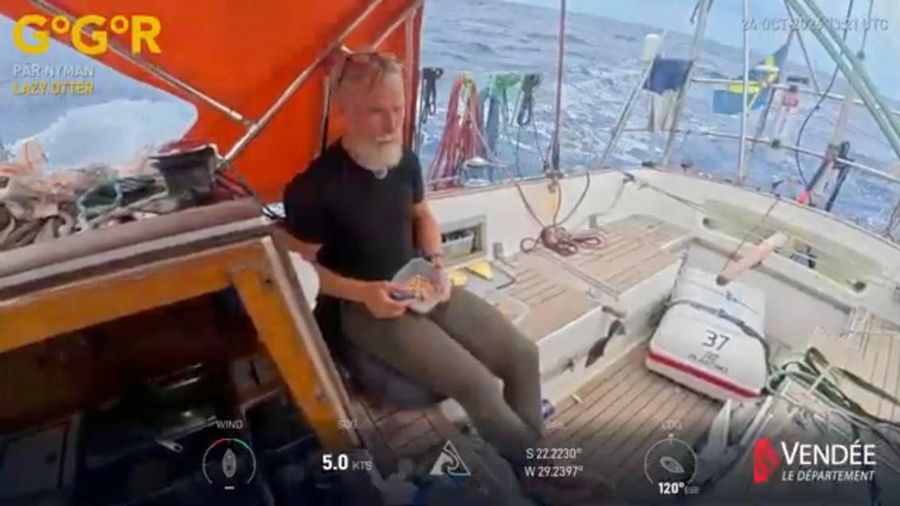 Pär Nyman sitting in the cockpit of Lazy Otter under an orange dodger, holding a small bowl, with the Swedish flag and a rolling sea behind him