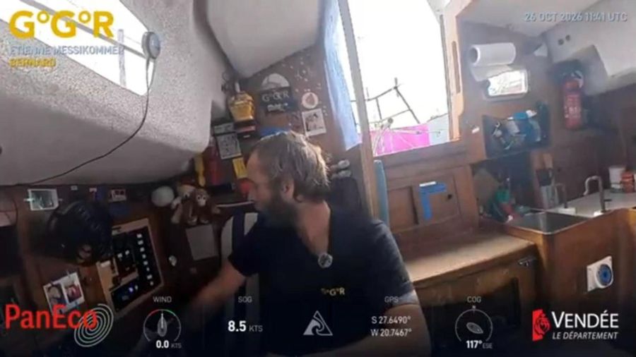 Etienne Messikommer, bearded and in a dark shirt, leaning over the instrument panel at the nav station of Bernard, with a bright window and galley sink behind him.