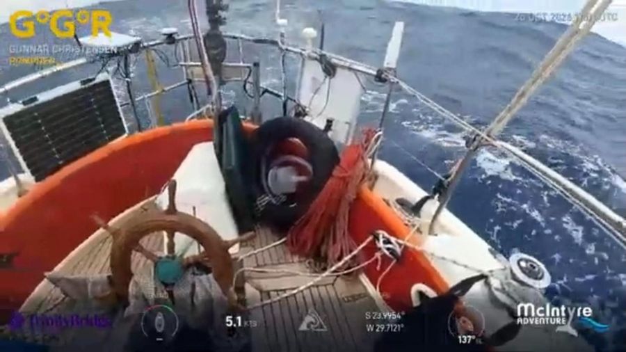 The cockpit of Pompoen from a camera inside the boat, with a wooden wheel, coiled orange line, a solar panel and a rough blue sea beyond the rail.