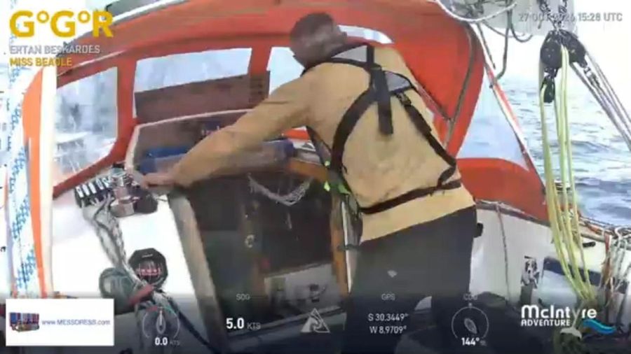 Ertan Beskardes in a tan top and harness leaning into the companionway of Miss Beagle, with a sail and blue sea beyond and the overlay showing 5.0 knots.