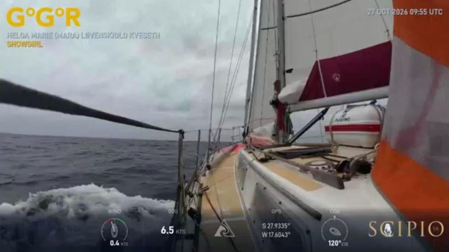The foredeck of Showgirl with a white life raft canister, a red sail bag and orange jib in the foreground, grey sea and sky and a breaking wave, the overlay showing 6.5 knots.