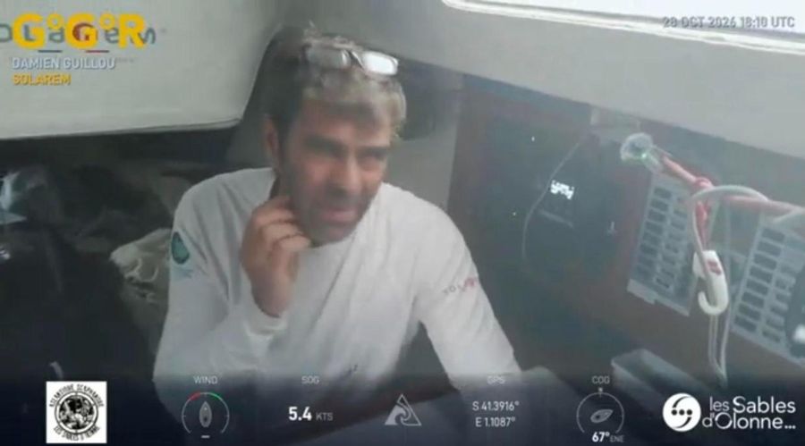 Damien Guillou in a white top with sunglasses on his head, sitting in the saloon of Solarem with a hand at his neck, the overlay showing 5.4 knots at 41 degrees south.