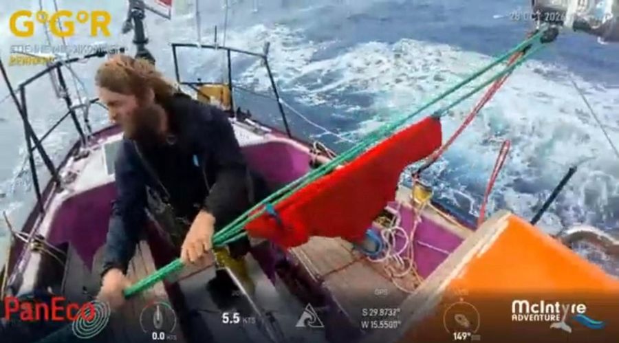 Etienne Messikommer, bearded and in dark oilskins, hauling on a green line beside a red sail on the pink and orange foredeck of Bernard, with white water alongside and the overlay showing 5.5 knots.