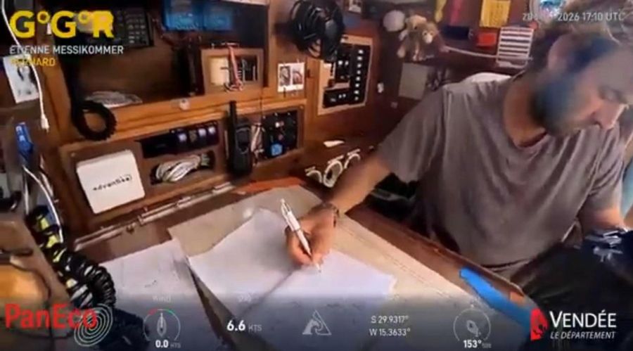 Etienne Messikommer in a grey T-shirt writing on a paper chart at the chart table of Bernard, with radios and switch panels behind him and the overlay showing 6.6 knots.