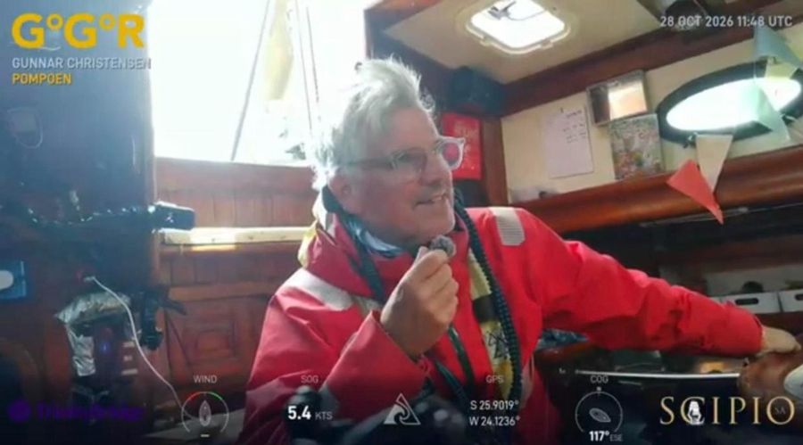 Gunnar Christensen in a red jacket with a lanyard around his neck, holding a radio handset in the cabin of Pompoen with a bright hatch behind him, the overlay showing 5.4 knots.