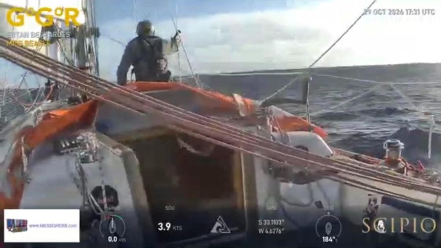 Ertan Beskardes, seen from behind in a grey jacket, standing on the foredeck of Miss Beagle with orange sails and lines in the foreground and a grey sea behind, the overlay showing 3.9 knots at 33 degrees south.