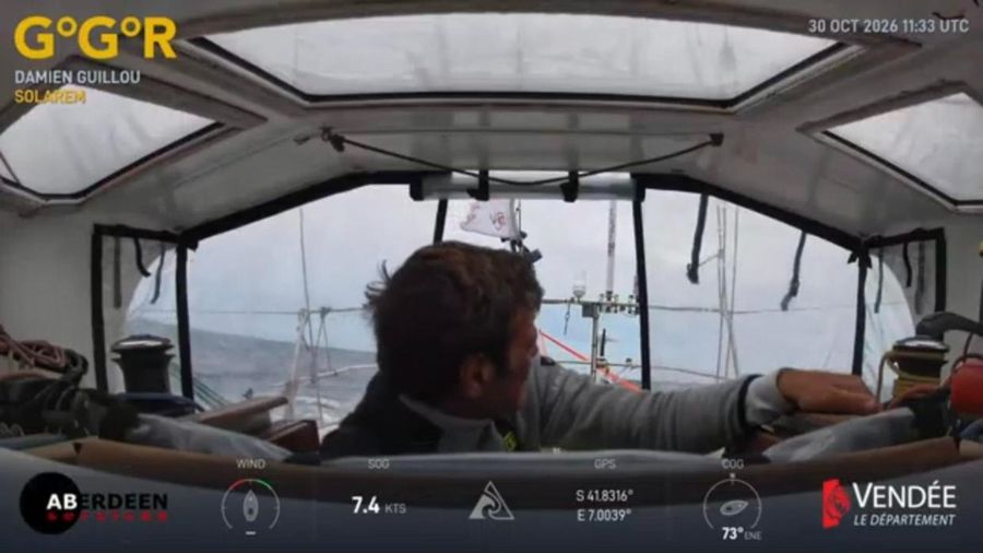 Damien Guillou seen from behind in the saloon of Solarem, looking out through the doghouse windows at a grey sea and the mast, the overlay showing 7.4 knots at 41.8 degrees south.