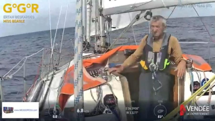 Ertan Beskardes in a lifejacket and tan top, standing at the companionway of Miss Beagle with an orange spray hood and white sail behind him and a grey sea beyond, the overlay showing 6.8 knots at 34 degrees south.