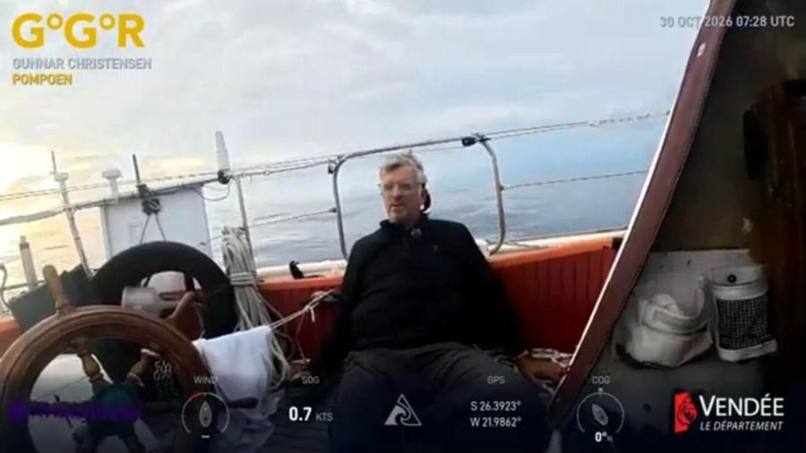Gunnar Christensen sitting in the cockpit of Pompoen beside the wooden wheel, with a flat grey sea and soft golden light behind him, the overlay showing 0.7 knots at 26 degrees south.