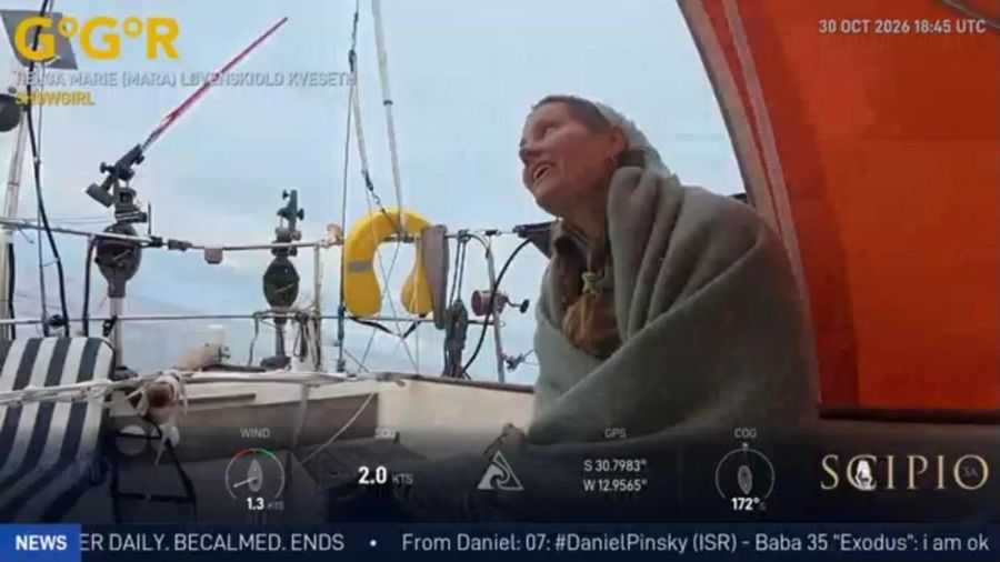 Mara Kveseth wrapped in a grey blanket in the cockpit of Showgirl, smiling and looking up at a pale sky, with a yellow lifebuoy and rigging behind her, the overlay showing 2.0 knots at 31 degrees south.